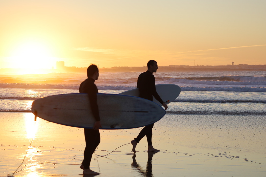 Duas pessoas com pranchas na praia, ao pôr do sol