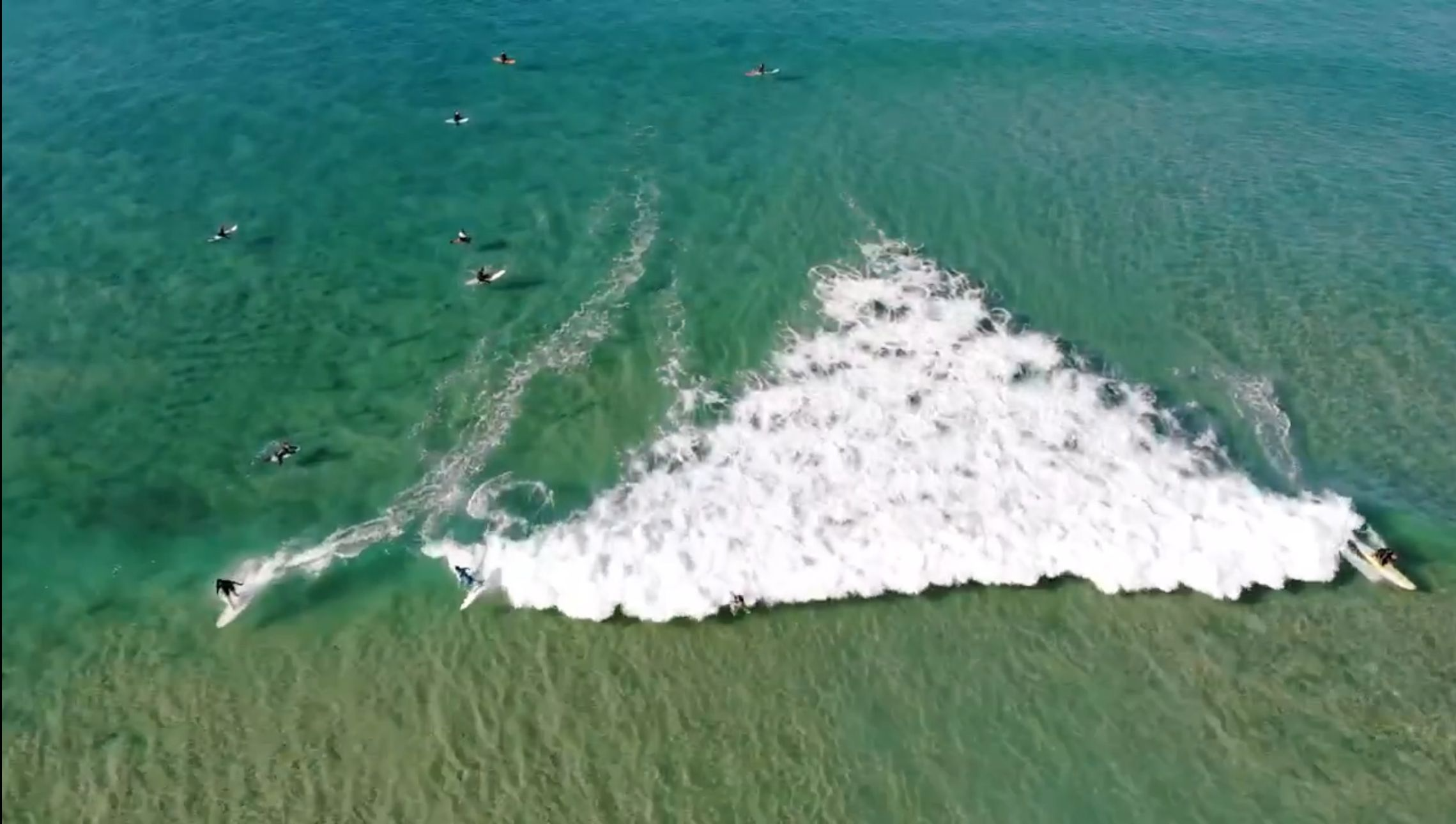 Vista aérea de surfistas numa onda, em água verde e transparente.